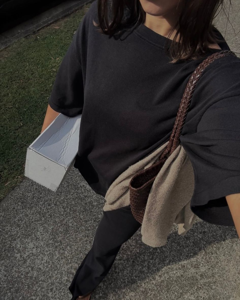 A person outdoors wearing an oversized black t-shirt and black pants, carrying a brown woven shoulder bag with a beige fabric pouch.
